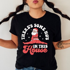 May include: Black t-shirt with a white graphic of Santa Claus in a split pose with the text "There's some ho's in this house".