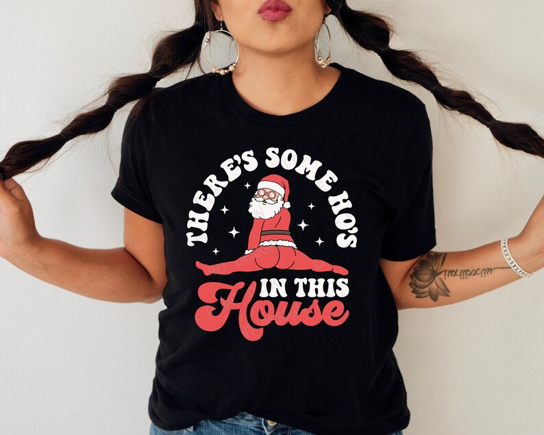 May include: Black t-shirt with a white graphic of Santa Claus in a split pose with the text "There's some ho's in this house".
