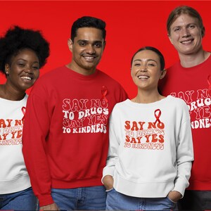 May include: Four people wearing red and white sweatshirts with the text "Say No To Drugs Say Yes To Kindness" in a retro font. Each sweatshirt features a red ribbon.