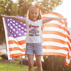 May include: A young person holding an American flag, wearing a white t-shirt with the text "Party in the USA" and blue denim shorts.