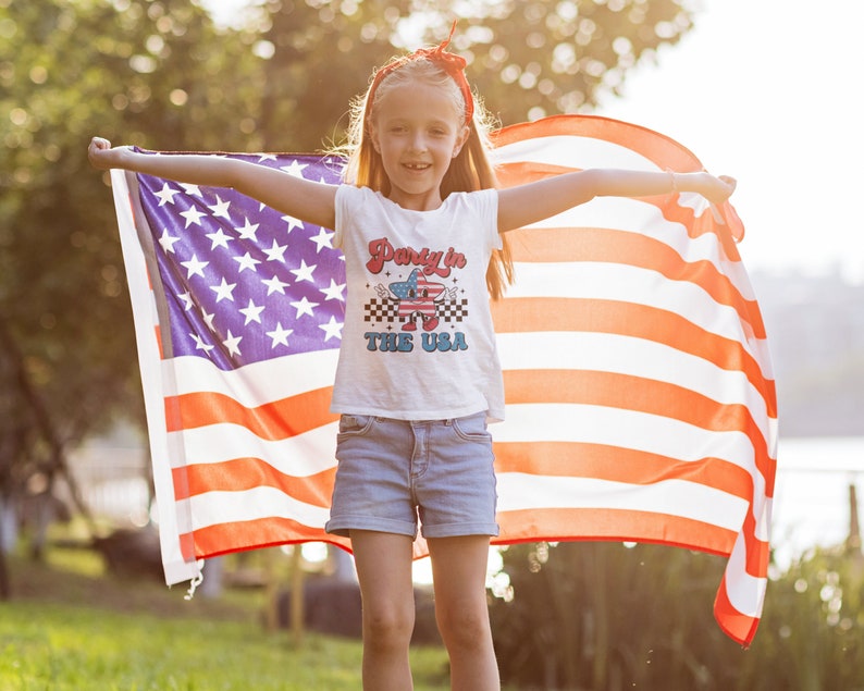 May include: A young person holding an American flag, wearing a white t-shirt with the text "Party in the USA" and blue denim shorts.