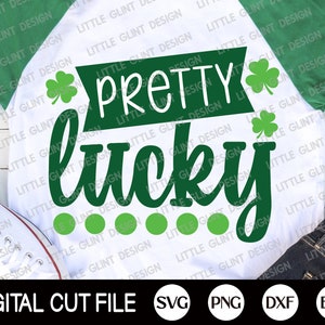 May include: A white t-shirt with green text that reads "Pretty Lucky" with four green shamrocks and a row of green dots. The shirt is laying on a wooden surface with a pair of white sneakers and blue jeans.