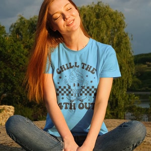 May include: A woman wearing a light blue t-shirt with a black and white graphic of an ice cream cone on a skateboard. The graphic says "Chill the 4th Out".