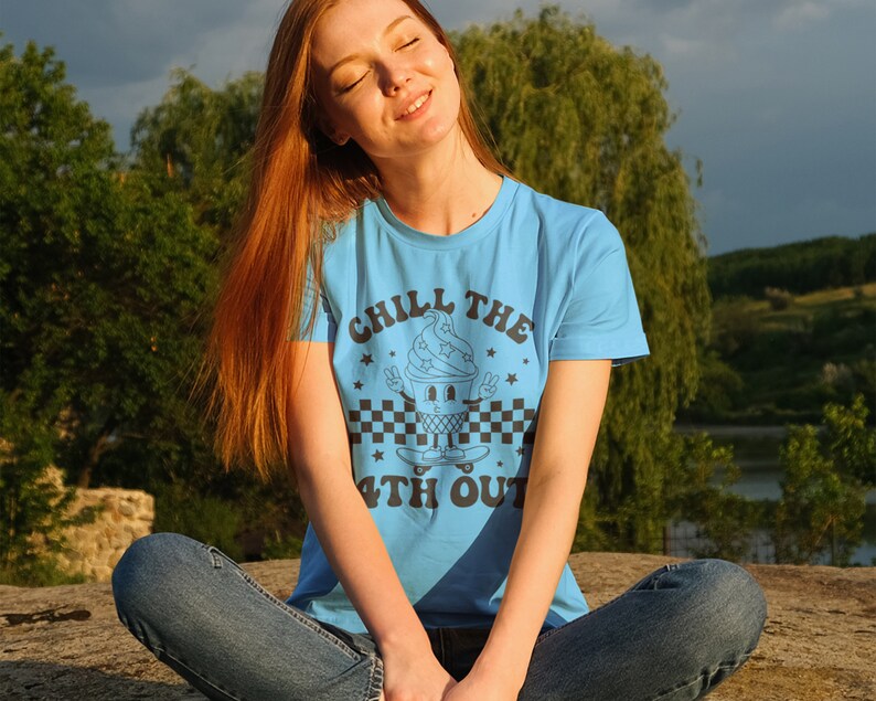 May include: A woman wearing a light blue t-shirt with a black and white graphic of an ice cream cone on a skateboard. The graphic says "Chill the 4th Out".