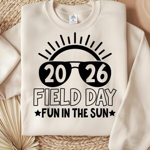 May include: Cream-colored sweatshirt with a black graphic. The graphic features the numbers "20 26" inside sunglasses, with the words "FIELD DAY" and "FUN IN THE SUN" below. The design includes a sun and stars.