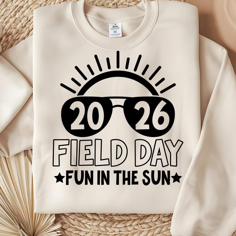 May include: Cream-colored sweatshirt with a black graphic. The graphic features the numbers "20 26" inside sunglasses, with the words "FIELD DAY" and "FUN IN THE SUN" below. The design includes a sun and stars.