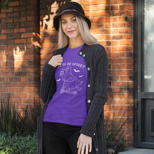May include: A purple t-shirt with the text "TO BE SPOOKY" and a white graphic of a ghost riding a scooter, with bats. The person is wearing a black hat and a dark gray cardigan.