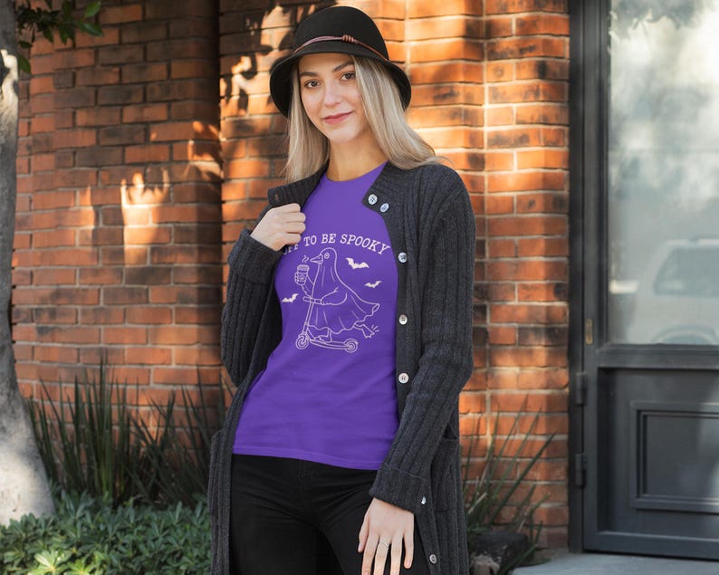 May include: A purple t-shirt with the text "TO BE SPOOKY" and a white graphic of a ghost riding a scooter, with bats. The person is wearing a black hat and a dark gray cardigan.