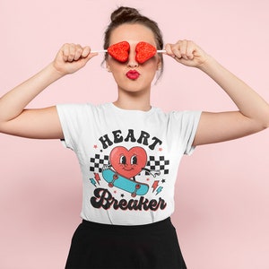 K&ouml;nnte beinhalten: Wei&szlig;es T-Shirt mit einem roten Herz-Motiv, das Skateboard f&auml;hrt, und dem Text "HEART BREAKER" in einer Retro-Schrift.