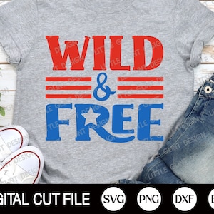 May include: A gray t-shirt with the text "Wild & Free" in red and blue lettering. The text is layered over three red stripes, reminiscent of the American flag. The shirt is laid out on a wooden surface with a pair of white sneakers and blue jeans.
