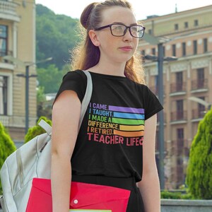 Puede incluir: Camiseta negra con el texto "I came, I taught, I made a difference, I retired, Teacher Life" en un dise&ntilde;o arco&iacute;ris. La camiseta la lleva una persona con gafas y una mochila.