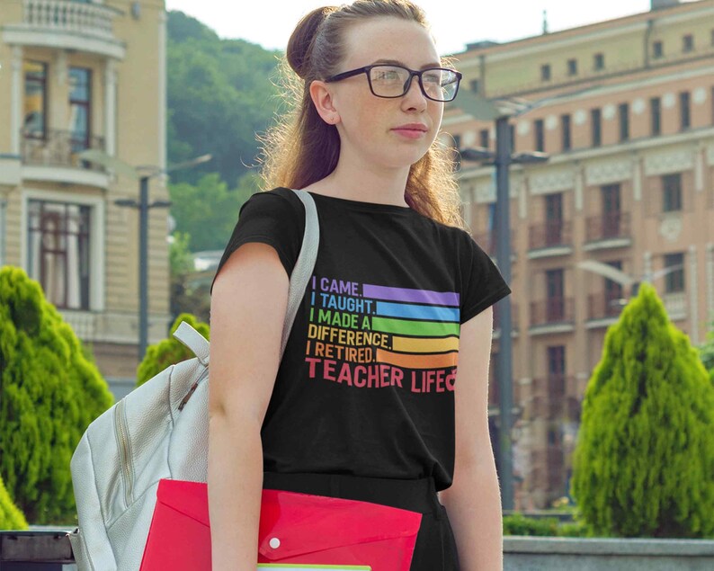 Puede incluir: Camiseta negra con el texto "I came, I taught, I made a difference, I retired, Teacher Life" en un dise&ntilde;o arco&iacute;ris. La camiseta la lleva una persona con gafas y una mochila.