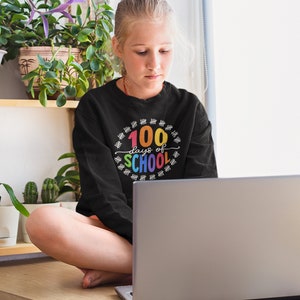 K&ouml;nnte beinhalten: Eine junge Person tr&auml;gt ein schwarzes Sweatshirt mit dem Text "100 Tage Schule" in Regenbogenfarben und einem Kreis aus Z&auml;hlstrichen. Die Person sitzt vor einem Laptop.