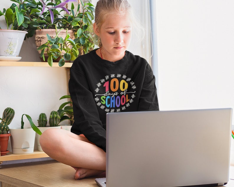 K&ouml;nnte beinhalten: Eine junge Person tr&auml;gt ein schwarzes Sweatshirt mit dem Text "100 Tage Schule" in Regenbogenfarben und einem Kreis aus Z&auml;hlstrichen. Die Person sitzt vor einem Laptop.