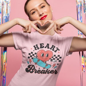K&ouml;nnte beinhalten: Ein pinkfarbenes T-Shirt mit einem Grafikdruck eines Cartoon-Herzens auf einem Skateboard. Das Herz hat ein l&auml;chelndes Gesicht und tr&auml;gt einen blauen Hut. Der Text "HEART BREAKER" ist in einer Retro-Schrift unter dem Grafikdruck gedruckt.