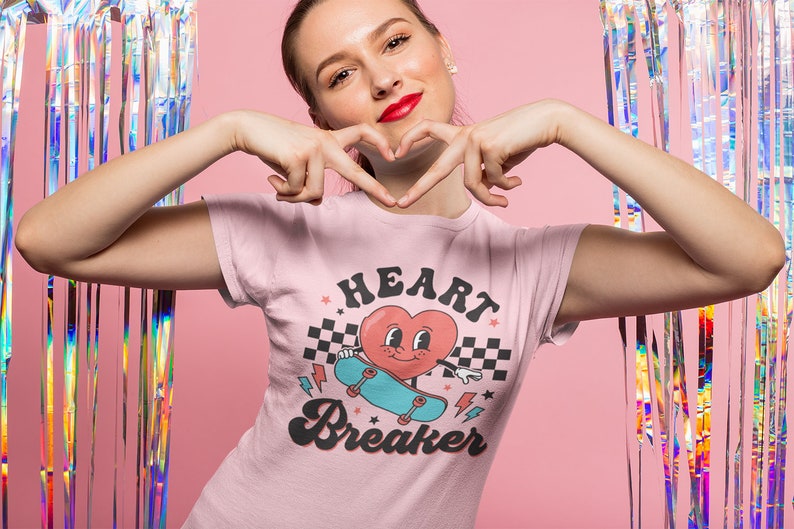 K&ouml;nnte beinhalten: Ein pinkfarbenes T-Shirt mit einem Grafikdruck eines Cartoon-Herzens auf einem Skateboard. Das Herz hat ein l&auml;chelndes Gesicht und tr&auml;gt einen blauen Hut. Der Text "HEART BREAKER" ist in einer Retro-Schrift unter dem Grafikdruck gedruckt.