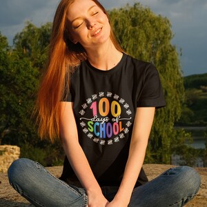 K&ouml;nnte beinhalten: Eine Frau tr&auml;gt ein schwarzes T-Shirt mit dem Text "100 Tage Schule" in Regenbogenfarben. Der Text ist von einem Kreis aus Z&auml;hlstrichen umgeben.