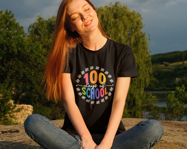 K&ouml;nnte beinhalten: Eine Frau tr&auml;gt ein schwarzes T-Shirt mit dem Text "100 Tage Schule" in Regenbogenfarben. Der Text ist von einem Kreis aus Z&auml;hlstrichen umgeben.