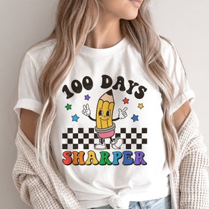 May include: White t-shirt with a colorful graphic of a pencil with a smiling face and the text "100 Days Sharper".