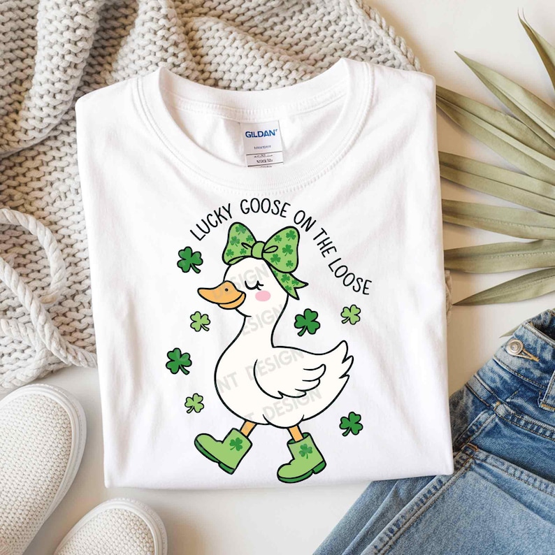 May include: White t-shirt with a cartoon goose design. The goose wears a green bow, boots, and is surrounded by shamrocks. The text "LUCKY GOOSE ON THE LOOSE" is printed above the goose in a circular pattern. The shirt is on a white surface.