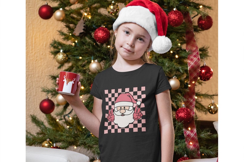 May include: A black t-shirt with a pink and white checkered pattern featuring a cartoon Santa Claus with a red hat and beard, making a peace sign with both hands. The text "Peace" is written in white letters on the shirt.