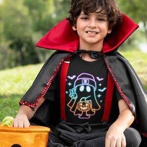 May include: A child wearing a black t-shirt with a rainbow ghost holding a jack-o-lantern and the text "Peace Out Ghost" printed on it. The child is also wearing a red and black cape.