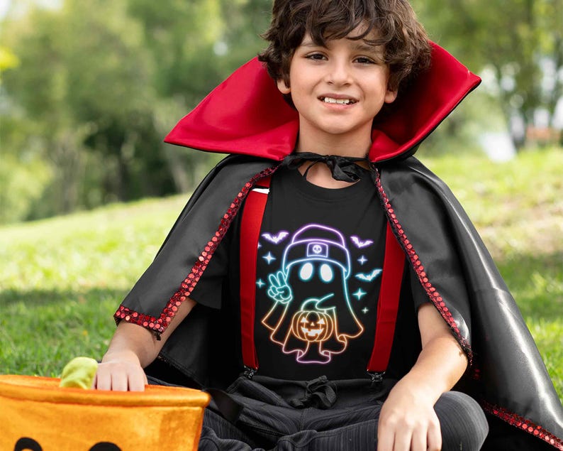 May include: A child wearing a black t-shirt with a rainbow ghost holding a jack-o-lantern and the text "Peace Out Ghost" printed on it. The child is also wearing a red and black cape.
