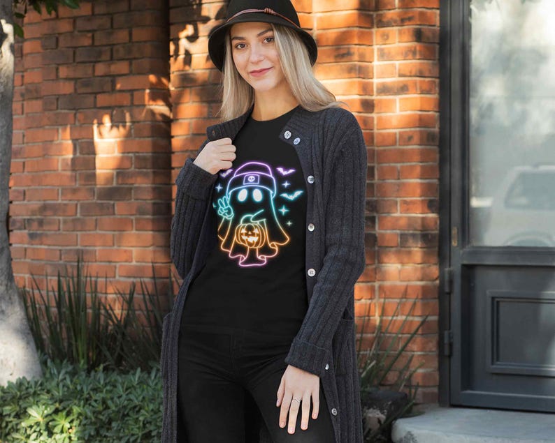 May include: A black t-shirt featuring a rainbow outline of a ghost holding a pumpkin. The ghost is wearing a hat. The design's background includes bats and stars.