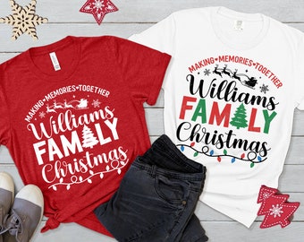 Può includere: Due magliette di Natale rosse e bianche con il testo "Making Memories Together Williams Family Christmas". Le magliette sono disposte su una superficie di legno con un paio di jeans e un ornamento di albero di Natale rosso.