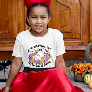 May include: A white t-shirt with the text "JUST WAITING FOR HALLOWEEN" and a cartoon ghost floating in a pool float. The ghost is holding a drink. The girl is wearing a red headband and a red skirt.