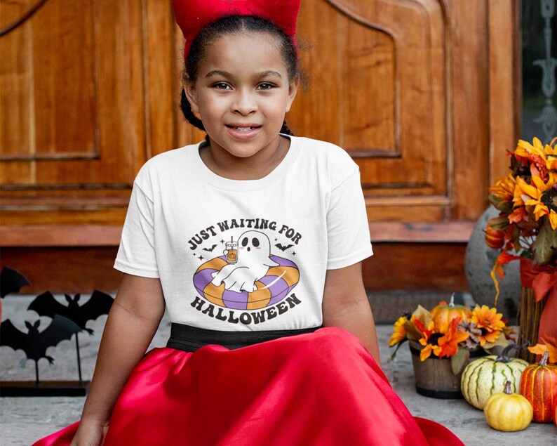 May include: A white t-shirt with the text "JUST WAITING FOR HALLOWEEN" and a cartoon ghost floating in a pool float. The ghost is holding a drink. The girl is wearing a red headband and a red skirt.