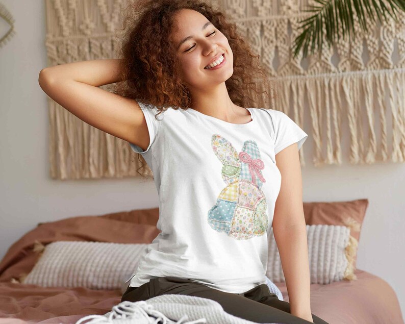 May include: White t-shirt featuring a patchwork bunny design with pastel colors and a pink bow. The bunny is made of various patterned fabrics, including floral and gingham prints. The shirt is short-sleeved and has a crew neck.