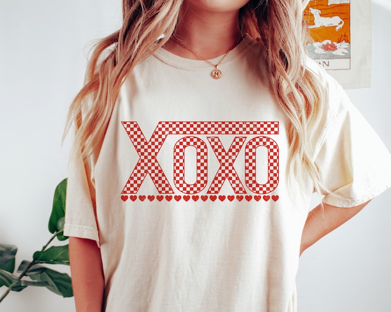 Pu&ograve; includere: Una maglietta bianca con una grafica "XOXO" a scacchi rossi e bianchi e una fila di cuori rossi sotto il testo.