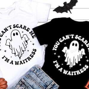 May include: Two white t-shirts with black text and a ghost graphic. The text reads "You can't scare me I'm a waitress".
