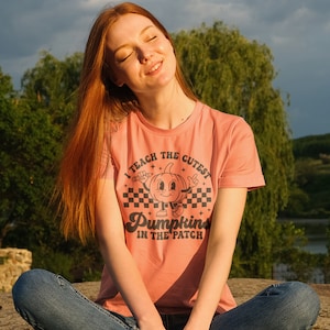 May include: A woman wearing a peach colored t-shirt with a black graphic that says "I teach the cutest pumpkins in the patch". The graphic features a cartoon pumpkin with a smiling face and arms.