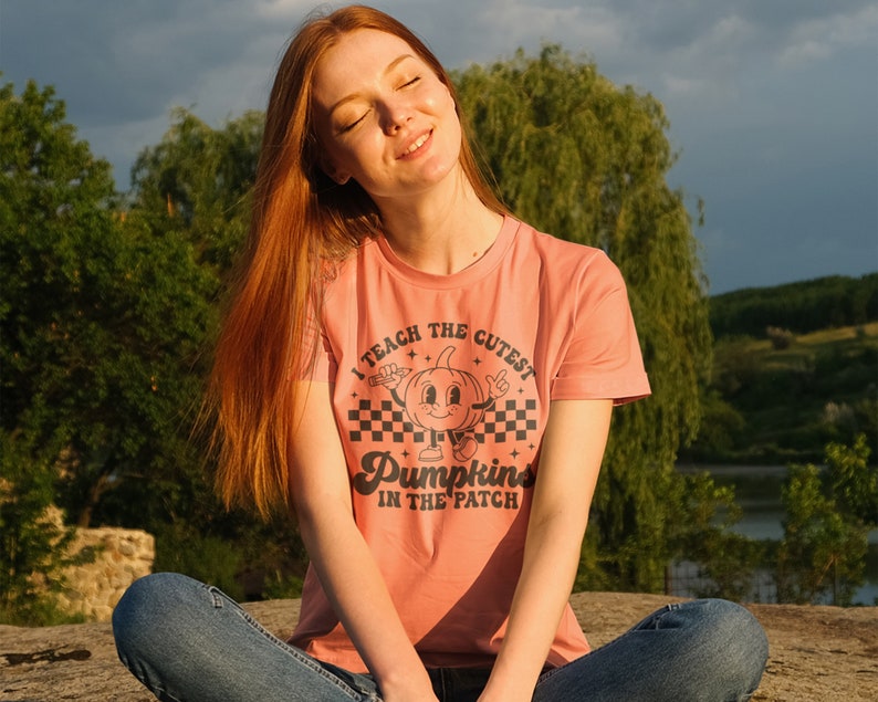 May include: A woman wearing a peach colored t-shirt with a black graphic that says "I teach the cutest pumpkins in the patch". The graphic features a cartoon pumpkin with a smiling face and arms.
