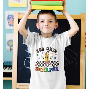 May include: A white t-shirt with a yellow pencil character wearing a red and white checkered hat. The text "100 Days Sharper" is printed on the shirt in rainbow colors.
