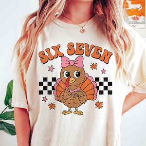 Pu&ograve; includere: T-shirt color crema con un tacchino dei cartoni animati con un fiocco rosa, piume arancioni e marroni e la scritta "SIX SEVEN" in arancione. Il design include stelle, foglie autunnali e motivi a scacchiera.
