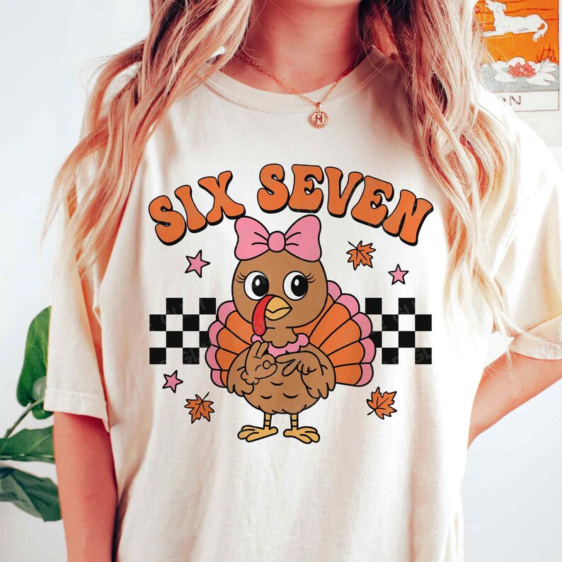 Pu&ograve; includere: T-shirt color crema con un tacchino dei cartoni animati con un fiocco rosa, piume arancioni e marroni e la scritta "SIX SEVEN" in arancione. Il design include stelle, foglie autunnali e motivi a scacchiera.