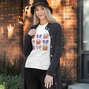 K&ouml;nnte beinhalten: Ein wei&szlig;es T-Shirt mit einem Halloween-Motiv, das mehrere orangefarbene und gelbe K&uuml;rbisse mit lila und rosa Schleifen zeigt. Die K&uuml;rbisse sind in einem sich wiederholenden Muster auf dem Shirt angeordnet.