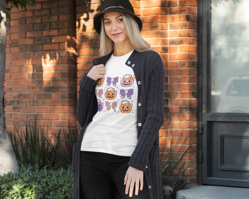 K&ouml;nnte beinhalten: Ein wei&szlig;es T-Shirt mit einem Halloween-Motiv, das mehrere orangefarbene und gelbe K&uuml;rbisse mit lila und rosa Schleifen zeigt. Die K&uuml;rbisse sind in einem sich wiederholenden Muster auf dem Shirt angeordnet.
