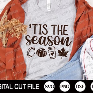 May include: A light grey t-shirt featuring the text "'Tis the Season" in brown script, with autumn-themed icons. The design includes a football, pumpkin, coffee cup, and maple leaf. The shirt is displayed with a rust-coloured jacket, jeans, and white trainers.
