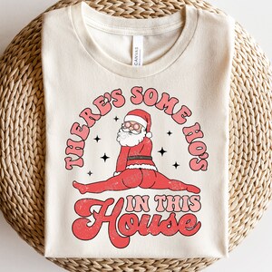 May include: A beige t-shirt with a red, white, and black graphic of Santa Claus in a split pose with the text "There's Some Ho's In This House".
