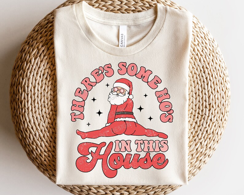 May include: A beige t-shirt with a red, white, and black graphic of Santa Claus in a split pose with the text "There's Some Ho's In This House".