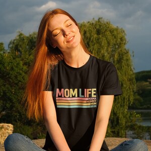 May include: A woman wearing a black t-shirt with the text "MOM LIFE" in rainbow colours and a striped design. The text "MAMA, MOMMY, MOM, BRUH" is printed below the striped design.