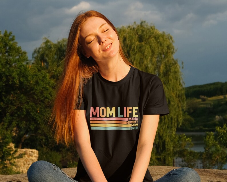 May include: A woman wearing a black t-shirt with the text "MOM LIFE" in rainbow colours and a striped design. The text "MAMA, MOMMY, MOM, BRUH" is printed below the striped design.