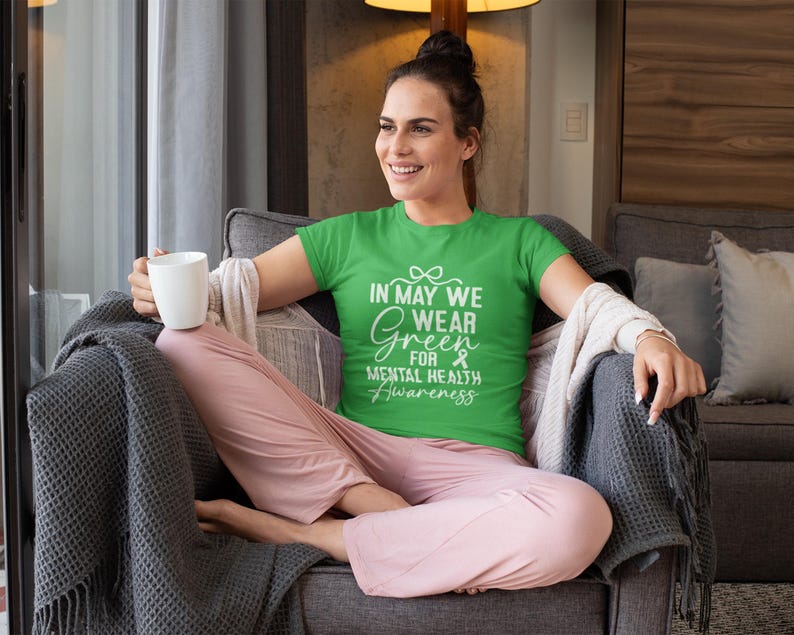 Op de afbeelding: Een groen t-shirt met de tekst "In May We Wear Green for Mental Health Awareness" in het wit. Het shirt wordt gedragen door een persoon die in een grijze fauteuil zit, een roze broek draagt en een witte mok vasthoudt.