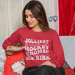 May include: A red sweatshirt with the text "Jolliest bunch of Hockey Moms this side of the Ice Rink" in white lettering. The person is holding a plate with a pastry. The background includes Christmas decorations.