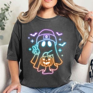 May include: A black t-shirt featuring a neon rainbow outline of a ghost wearing a hat and holding a jack-o'-lantern. The ghost has two eyes and a small mouth.
