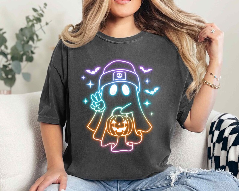 May include: A black t-shirt featuring a neon rainbow outline of a ghost wearing a hat and holding a jack-o'-lantern. The ghost has two eyes and a small mouth.